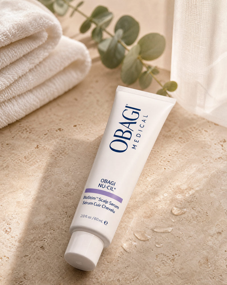 Obagi Nu-Cil BioStim Sérum Capilar recostado sobre travertino con gotas de agua