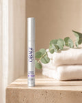 Obagi Nu-Cil Sérum Potenciador de Cejas de pie sobre repisa de travertino con toallas y eucalipto, vista frontal