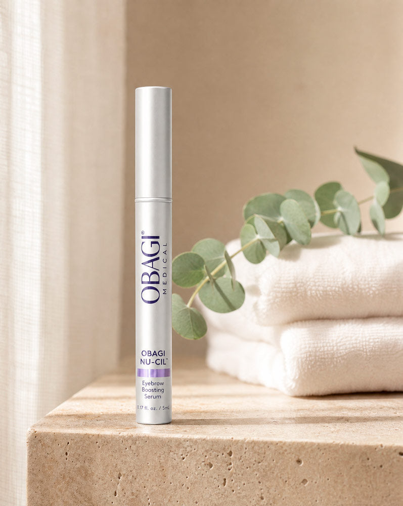 Obagi Nu-Cil Sérum Potenciador de Cejas de pie sobre repisa de travertino con toallas y eucalipto, vista frontal