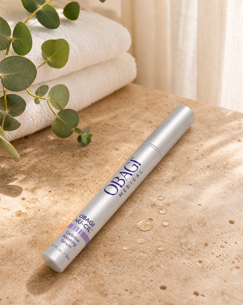 Obagi Nu-Cil Sérum Potenciador de Cejas recostado sobre travertino con gotas de agua