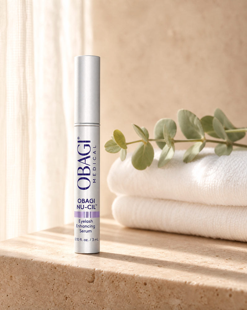 Obagi Nu-Cil Sérum Potenciador de Pestañas de pie sobre repisa de travertino con toallas y eucalipto, vista frontal
