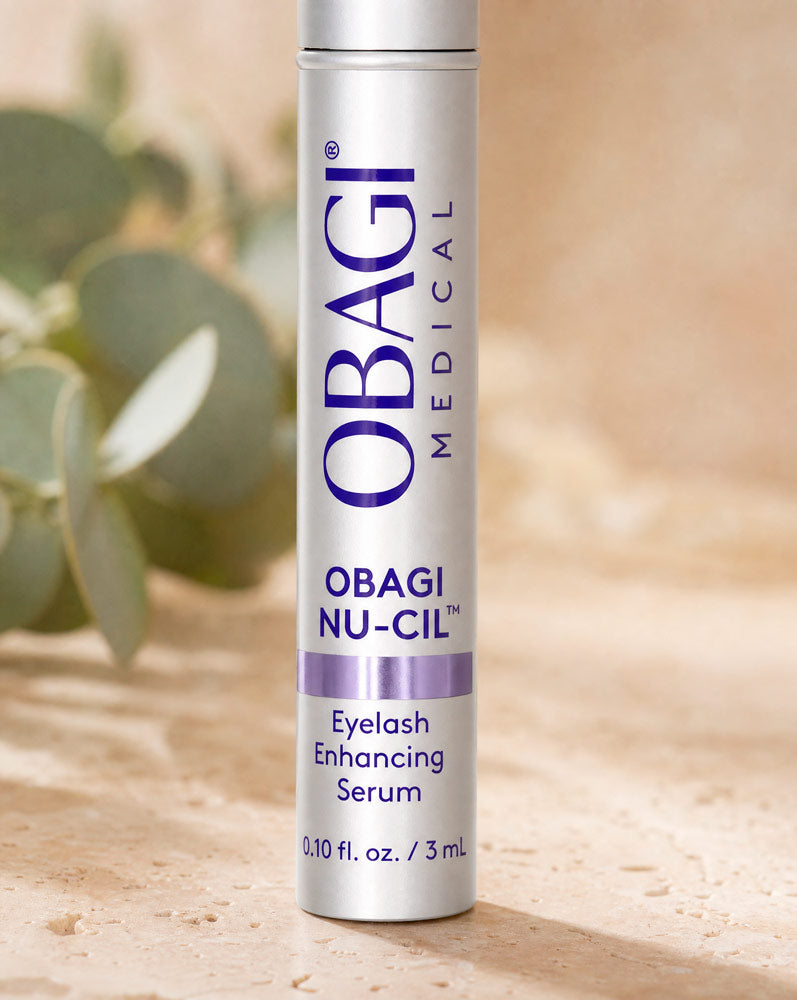 Obagi Nu-Cil Sérum Potenciador de Pestañas recostado sobre travertino con gotas de agua
